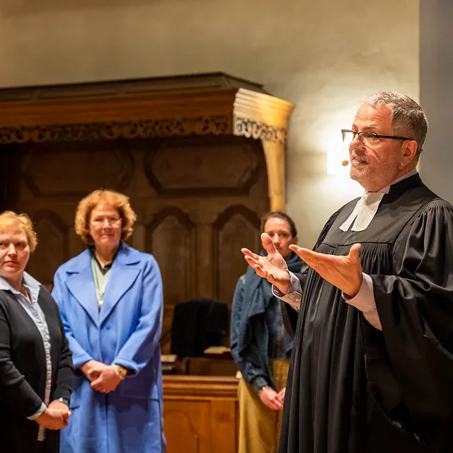 Mann im Talar spricht zu Frauen in Kirche.