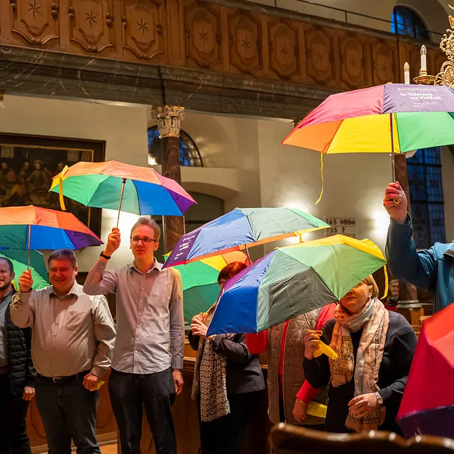 Mehrere Personen halten bunte Regenschirme in einer Kirche hoch.