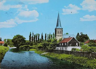 Historische Kirche an einem Fluss mit Bäumen und blauem Himmel.