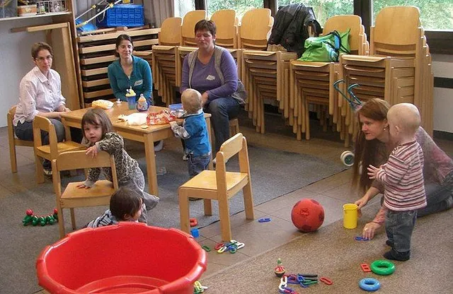 Kinder spielen in einem Raum, während Erwachsene an Tischen sitzen.