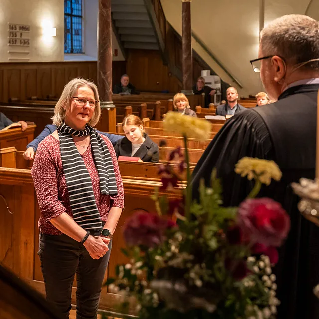 Eine Frau steht vor einem Prediger in einer Kirche, Blumen im Vordergrund.