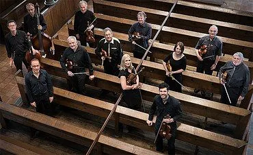 Ein Orchester von elf Musikern posiert in einer Kirche mit Streichinstrumenten.