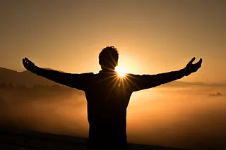 Silhouette einer Person mit ausgebreiteten Armen bei Sonnenaufgang über Nebel.