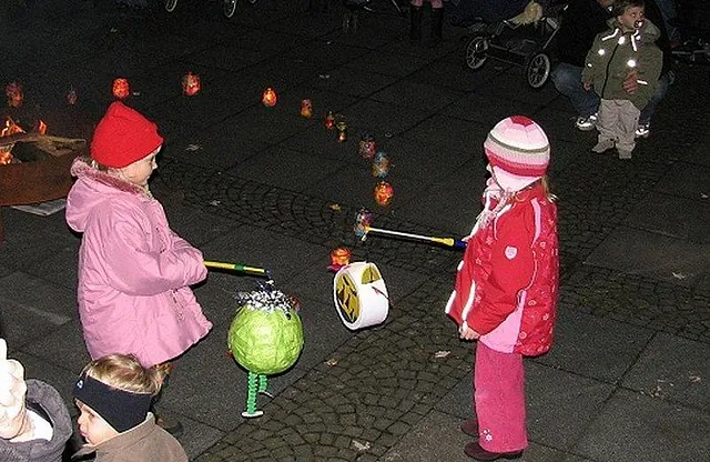 Zwei Kinder mit Laternen stehen nachts auf einem gepflasterten Bereich.